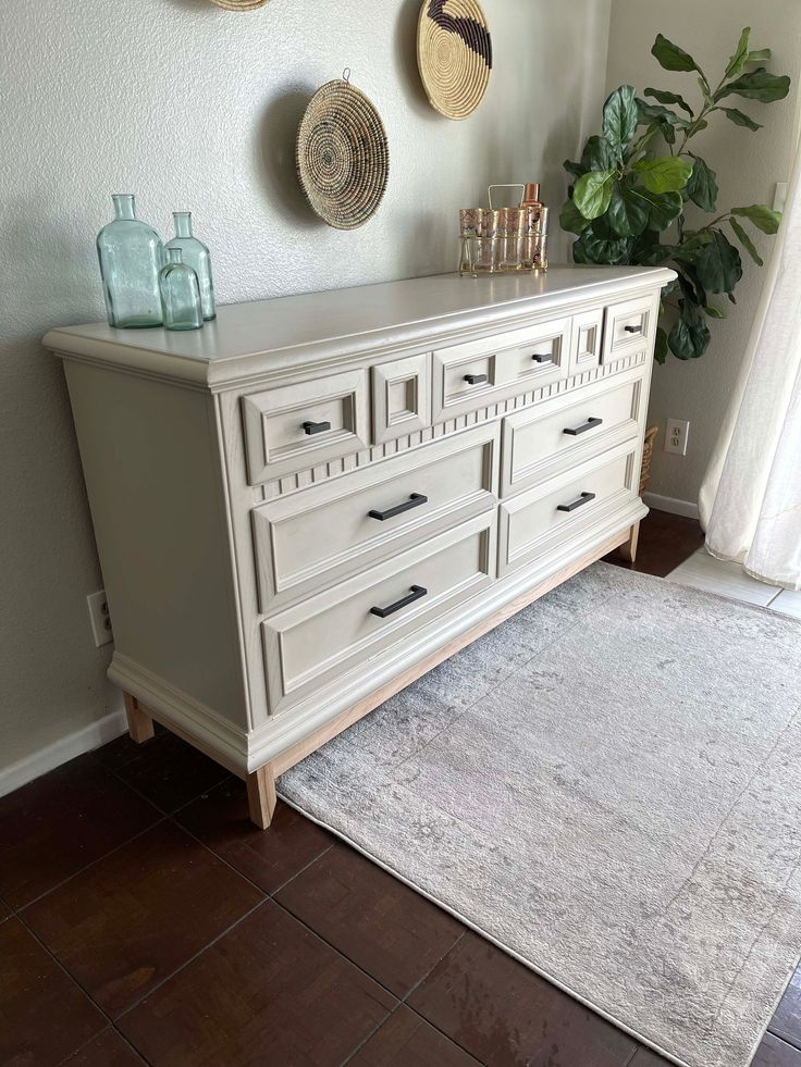 a white dresser sitting on top of a wooden floor next to a rug and potted plant