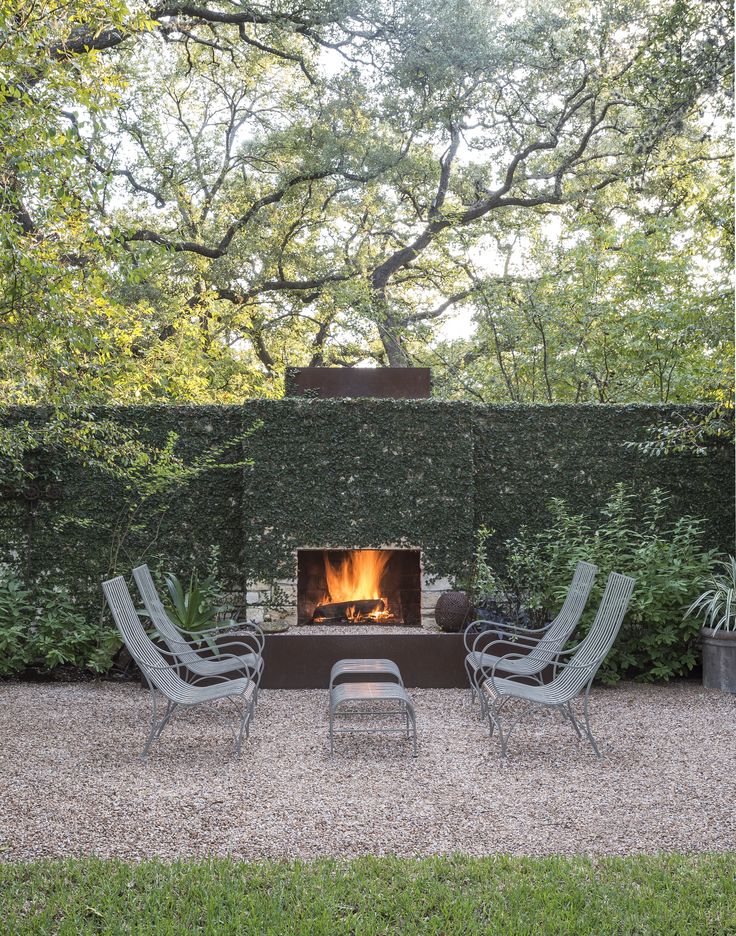 two lawn chairs sitting in front of a fire place with an outdoor fireplace surrounded by greenery