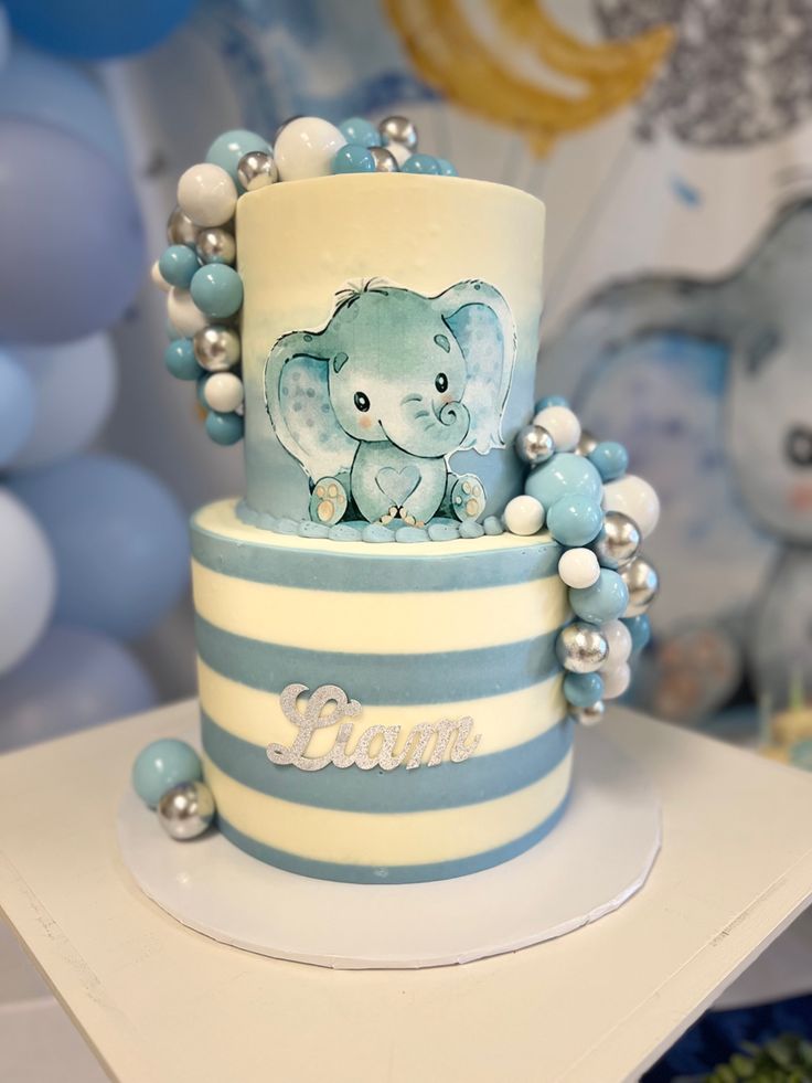 a blue and white striped cake with an elephant on it's top, surrounded by balloons