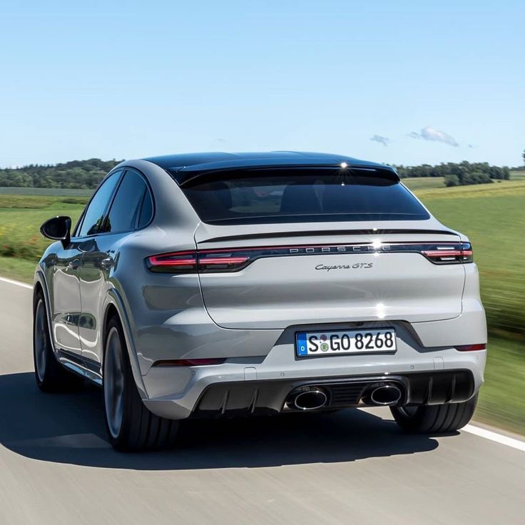 the rear end of a silver porsche cayenne driving on a road with grass and trees in the background