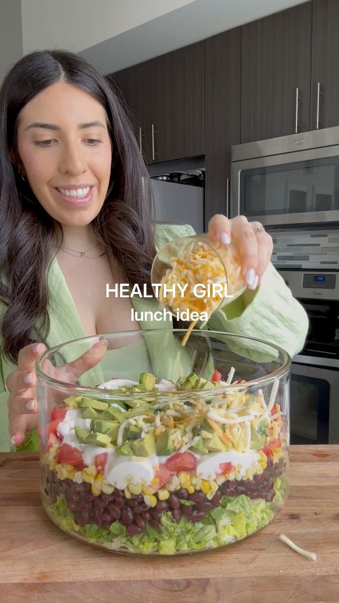 a woman is pouring dressing into a large glass bowl filled with vegetables and other ingredients