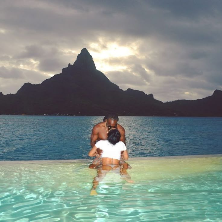 a man and woman are standing in the water by some mountains with blue sky behind them