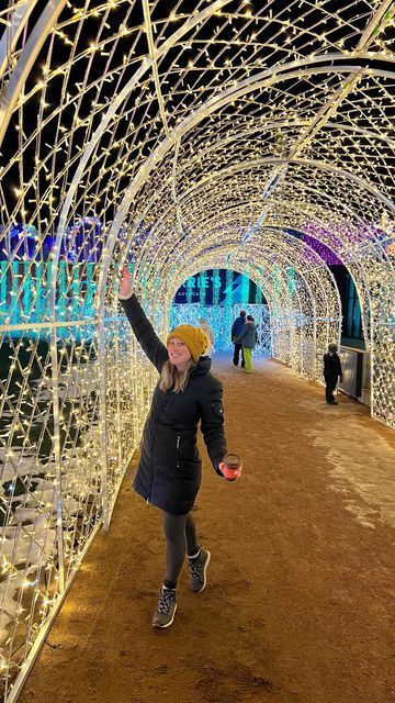 a woman is walking through a tunnel covered in christmas lights at the park with her arms up