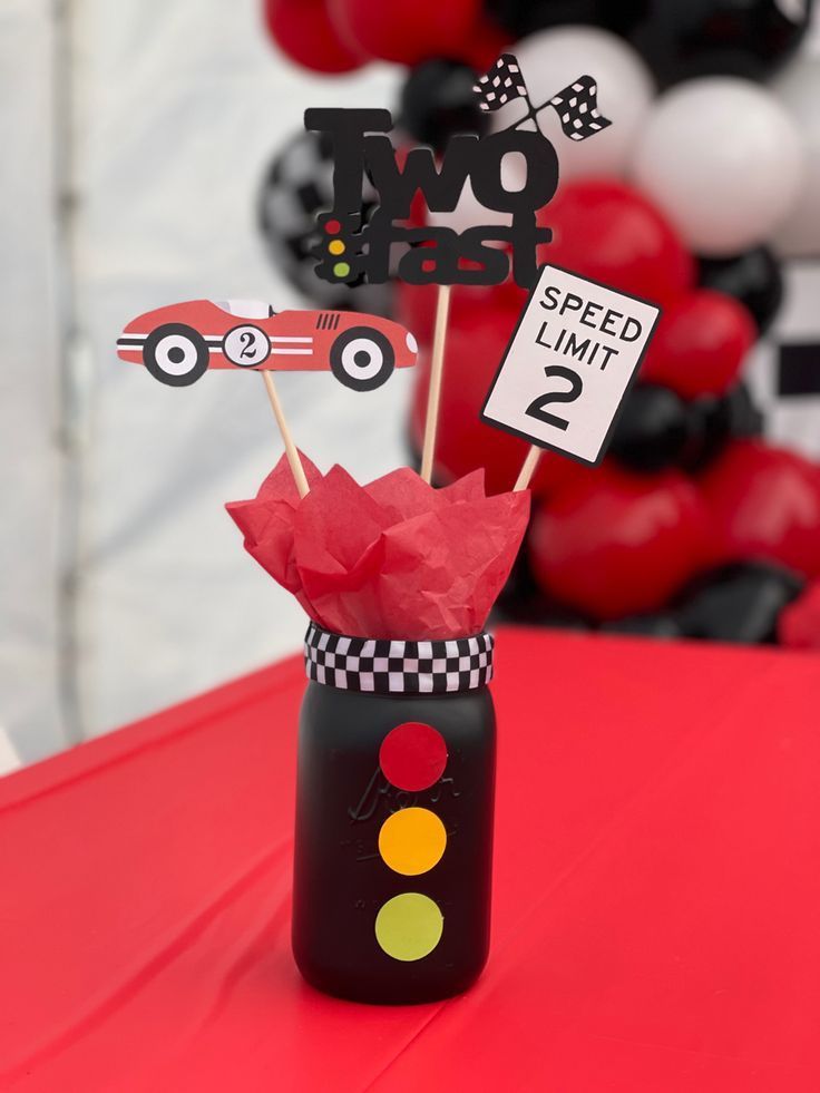 a red table topped with a cup filled with cake toppers and paper cars on sticks