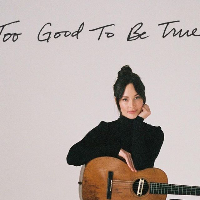 a woman sitting on top of a wooden chair holding a guitar in front of a white wall