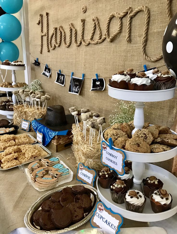 a table filled with lots of desserts on top of white trays next to blue and black balloons