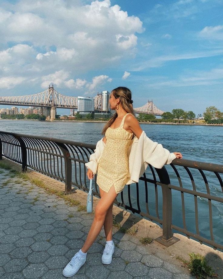 a woman in a short dress leaning on a fence by the water with a bridge in the background