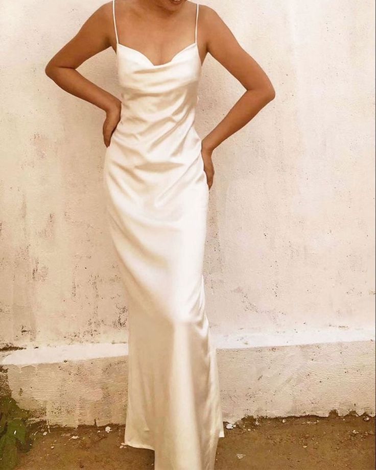 a woman standing in front of a white wall wearing a long dress with spaghetti straps