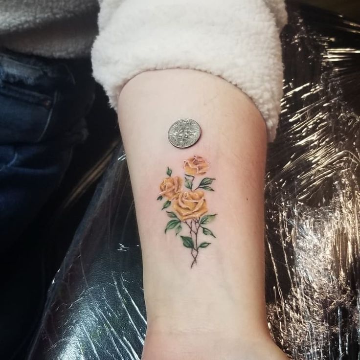 a small yellow rose tattoo on the left inner forearm and wrist with a coin sitting next to it