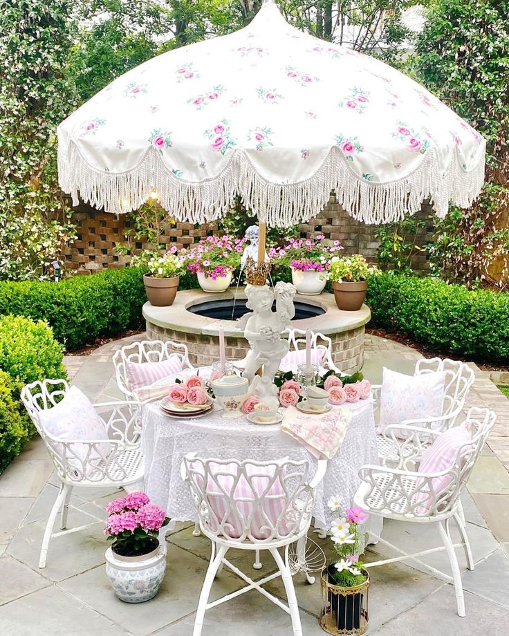 an outdoor table with white chairs and umbrella in the middle of a garden area, surrounded by potted plants