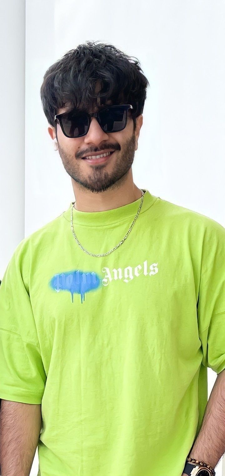 a man wearing sunglasses and a green shirt with the word angels on it, standing in front of a white wall