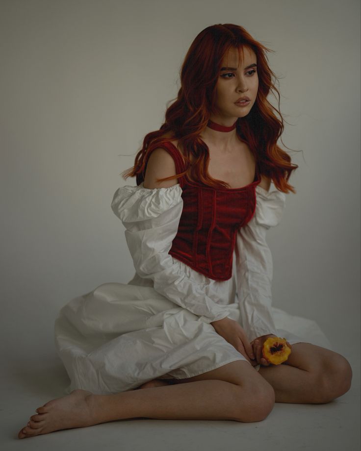 a woman with red hair sitting on the ground holding a banana in her hand and wearing a white dress