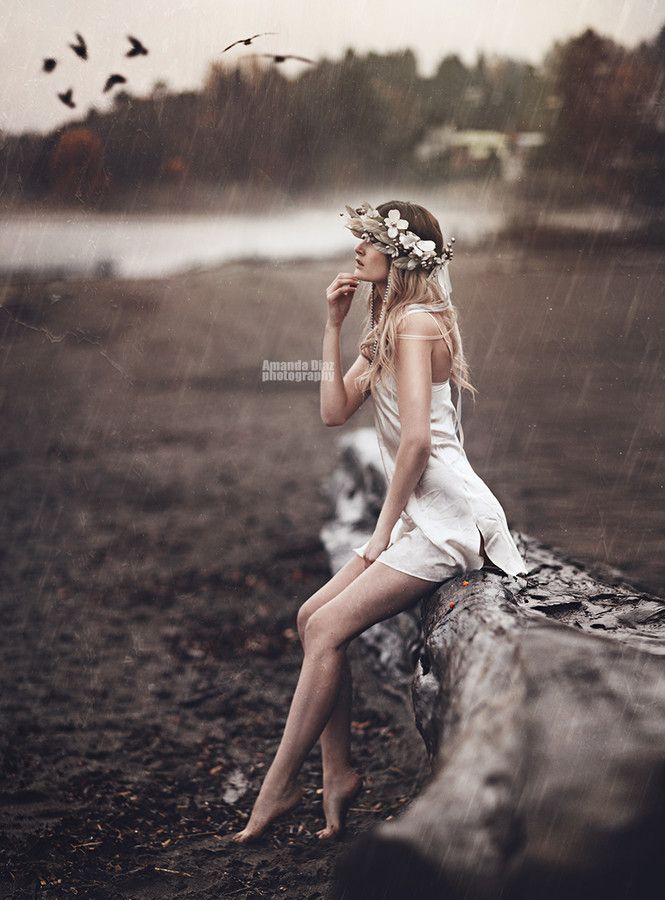 a woman sitting on a log in the rain with birds flying above her and trees behind her