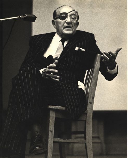 an old man sitting in a chair with his hand up to the side and holding a microphone