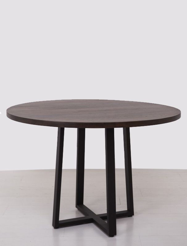 a round wooden table with black legs on a white floor in front of a gray wall