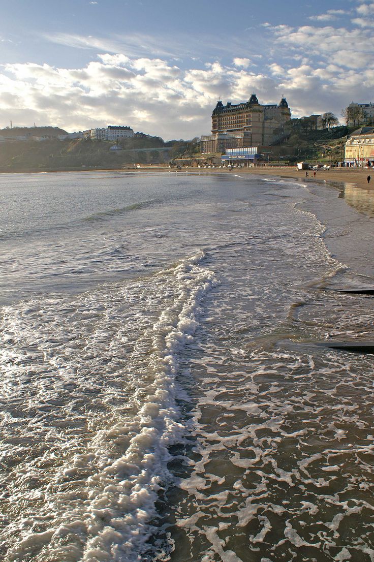 there is a wave coming in to the beach and buildings on the hill behind it