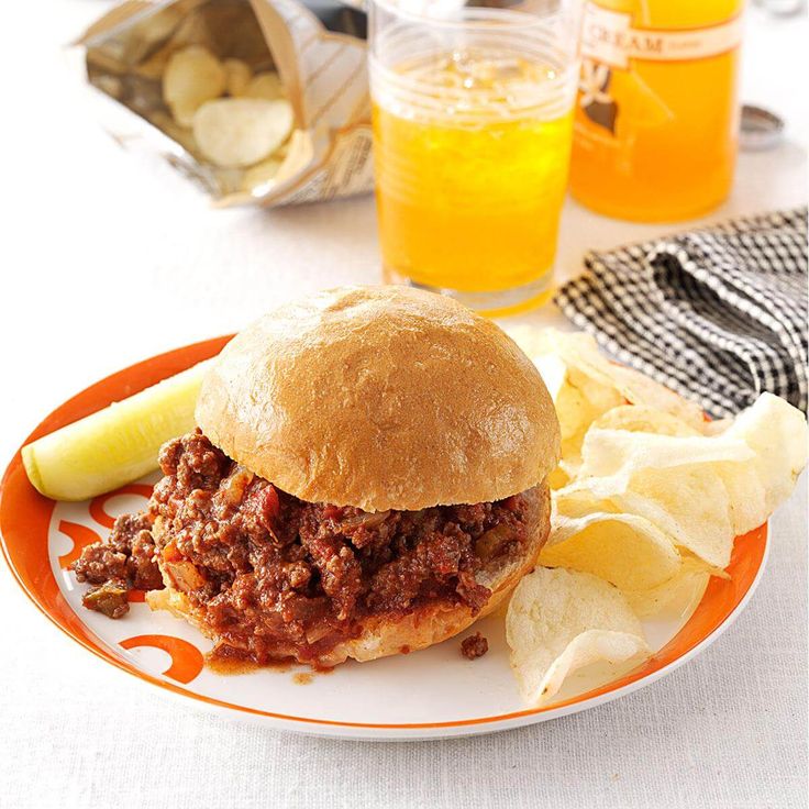 a sloppy joe sandwich on a plate with potato chips and orange juice in the background