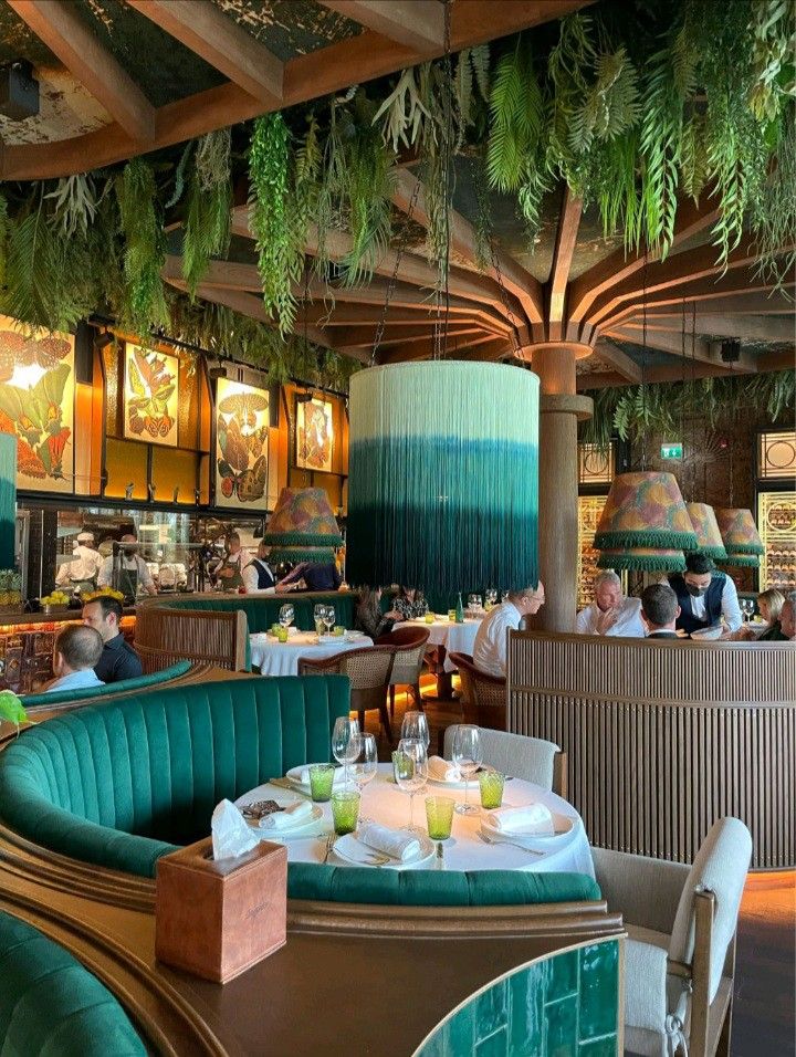 people sitting at tables in a restaurant with plants hanging from the ceiling and potted plants on the walls