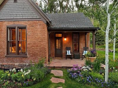 a small brick house sitting in the middle of a lush green yard with flowers and trees