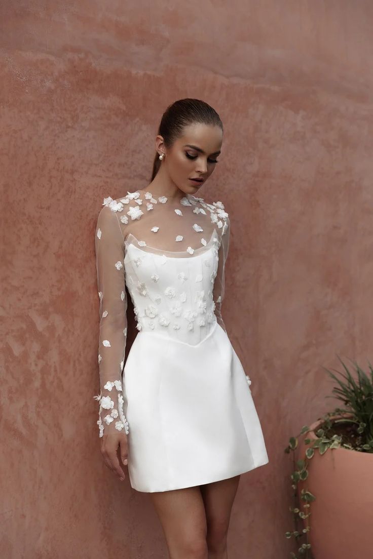 a woman in a short white dress standing next to a wall with flowers on it