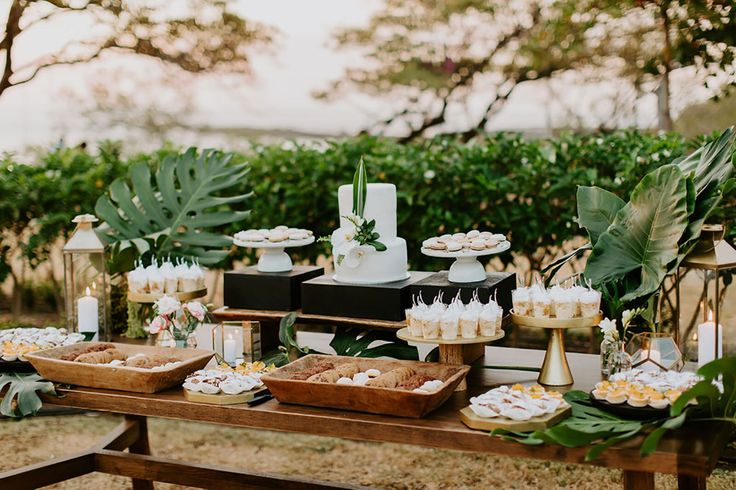 a table topped with lots of desserts on top of a wooden table covered in greenery