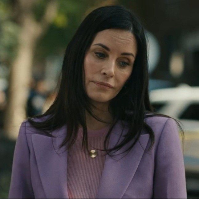 a woman in a purple suit and necklace looking at something on the ground with cars behind her