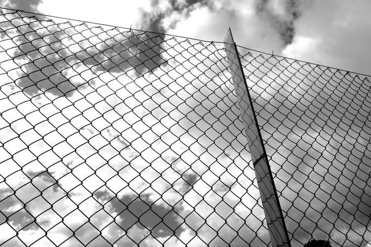 black and white photograph of clouds behind a chain link fence with the sky in the background