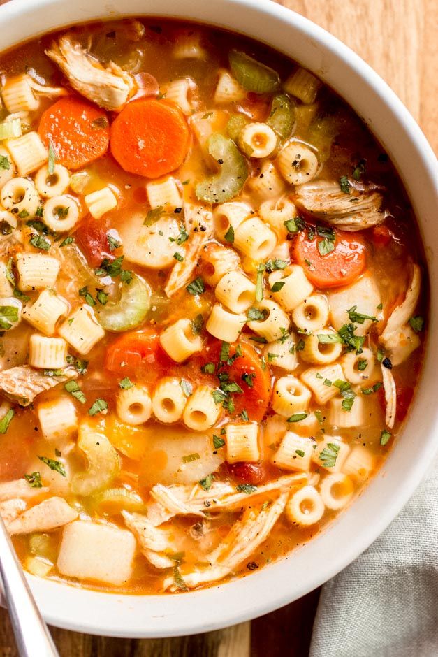 a white bowl filled with pasta and vegetable soup on top of a wooden table next to a spoon