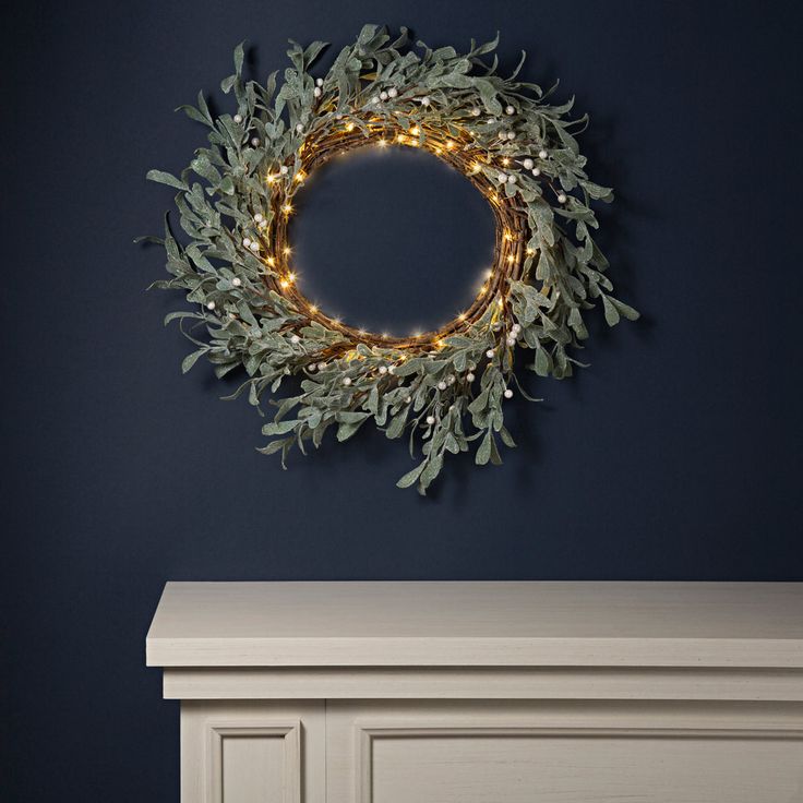 a wreath with lights hanging from it on the wall above a dresser in a dark blue room