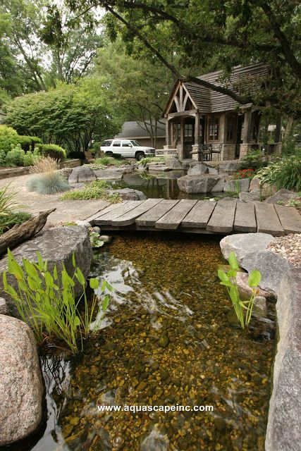 a small pond in the middle of a garden with rocks and plants around it, next to a house