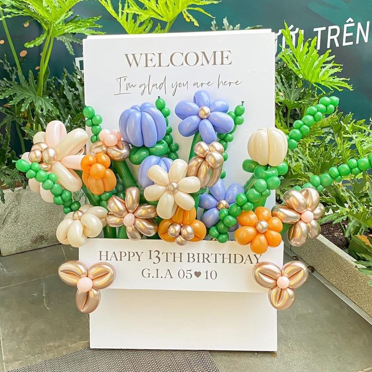 a welcome sign with balloons and flowers on it in front of some potted plants