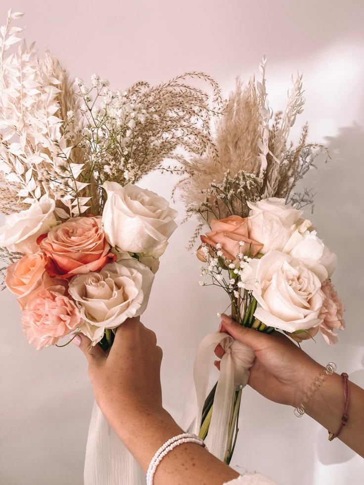 two hands holding bouquets of flowers in front of a white wall with dried grass