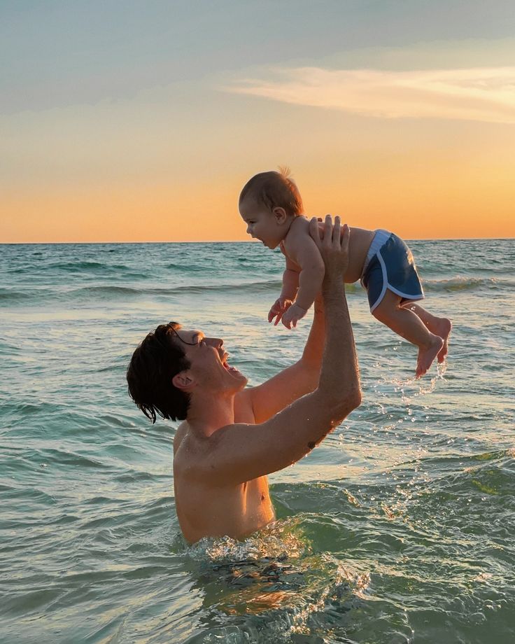 a man holding a baby up in the air while standing in the ocean at sunset