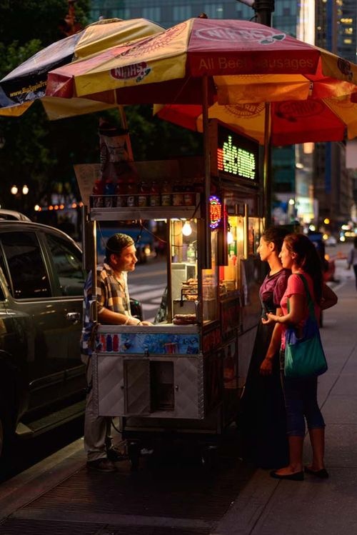 two people standing at a food cart on the sidewalk