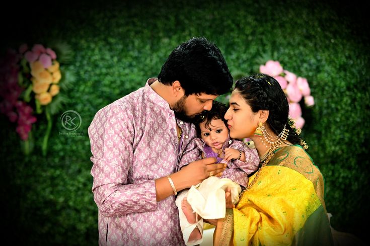 a man and woman holding a baby in front of green grass with flowers behind them