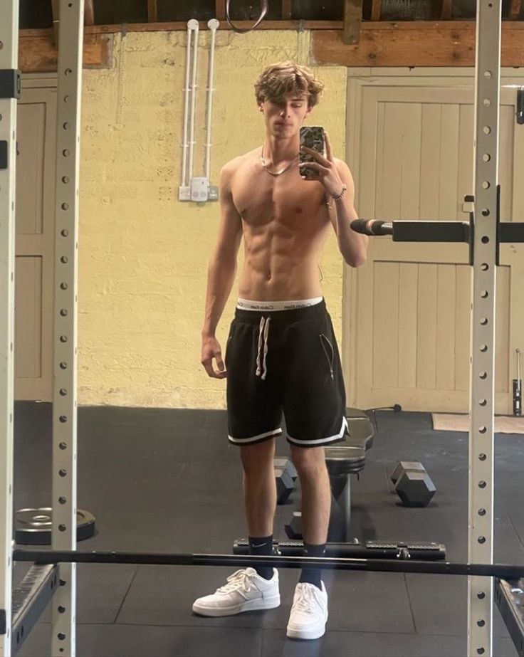 a shirtless man takes a selfie in the gym with his cell phone while wearing shorts