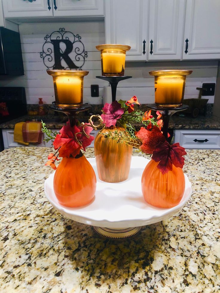 three orange vases on a white plate with flowers and candles in front of them