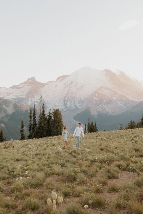 two people are walking in the grass with mountains in the backgrounds