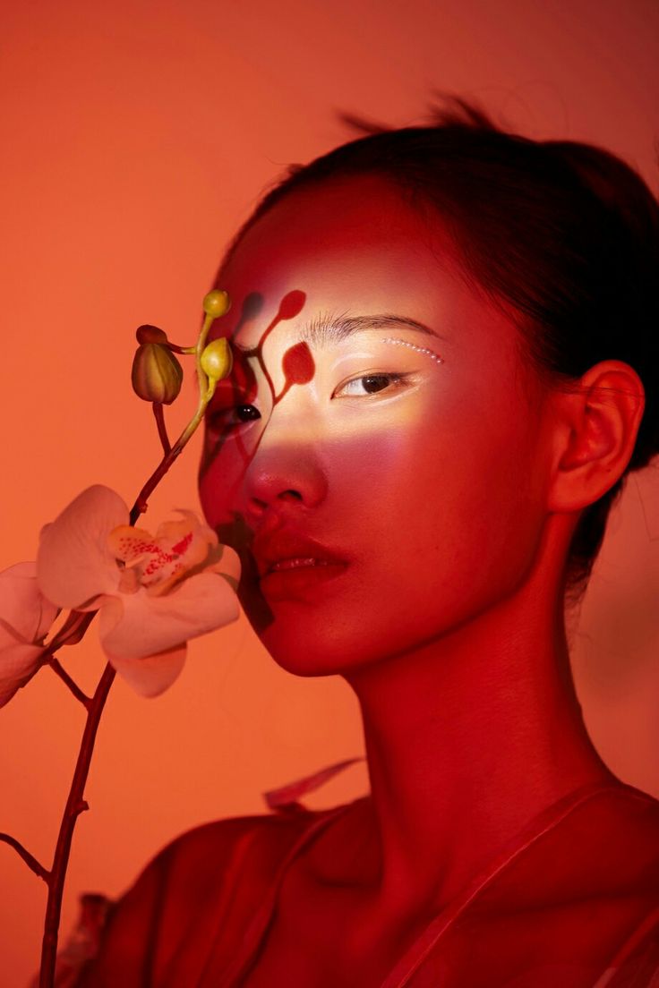 a woman with makeup on her face and flower in front of her face, against an orange background