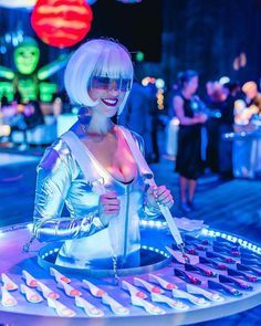 a woman dressed in silver holding a knife and standing next to a table with food on it