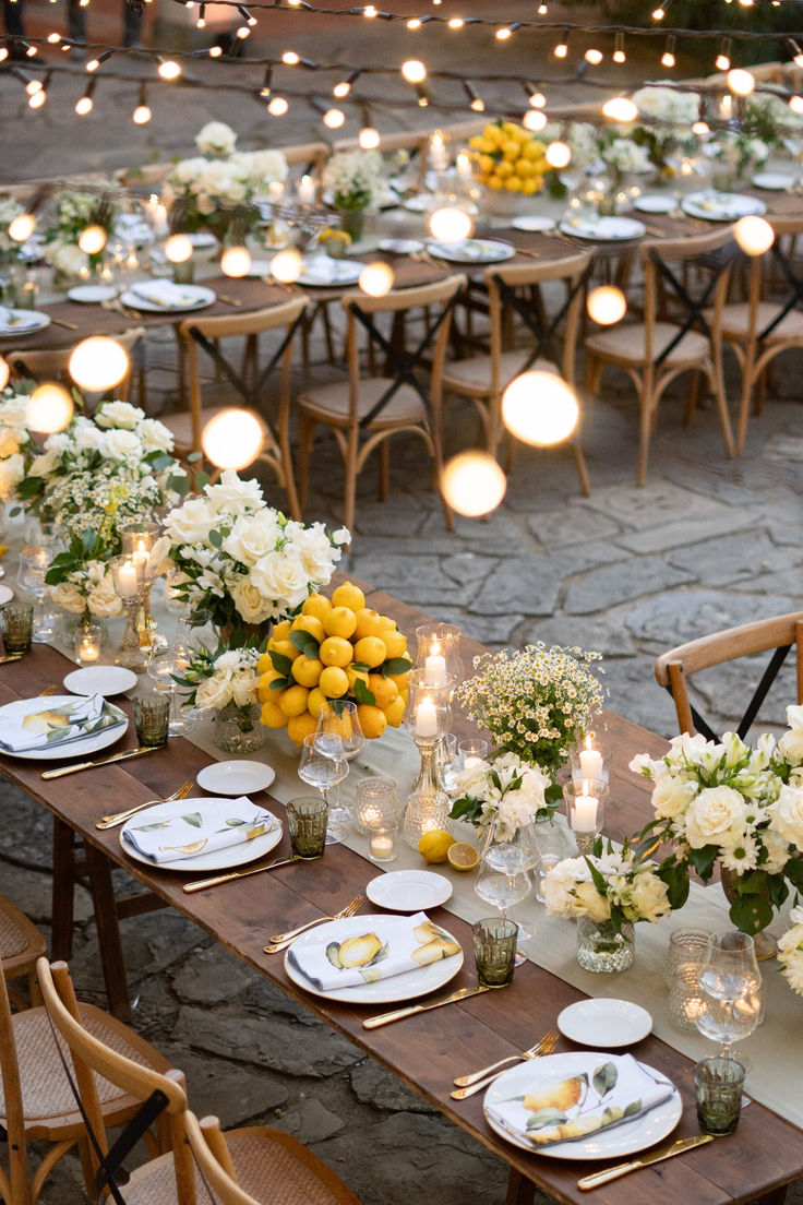 a long table is set with white and yellow floral centerpieces, candles, and plates