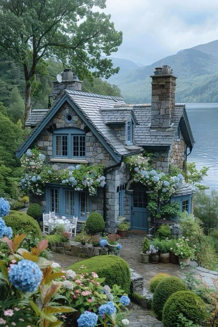 a stone house with blue shutters and flowers in the foreground, near a body of water