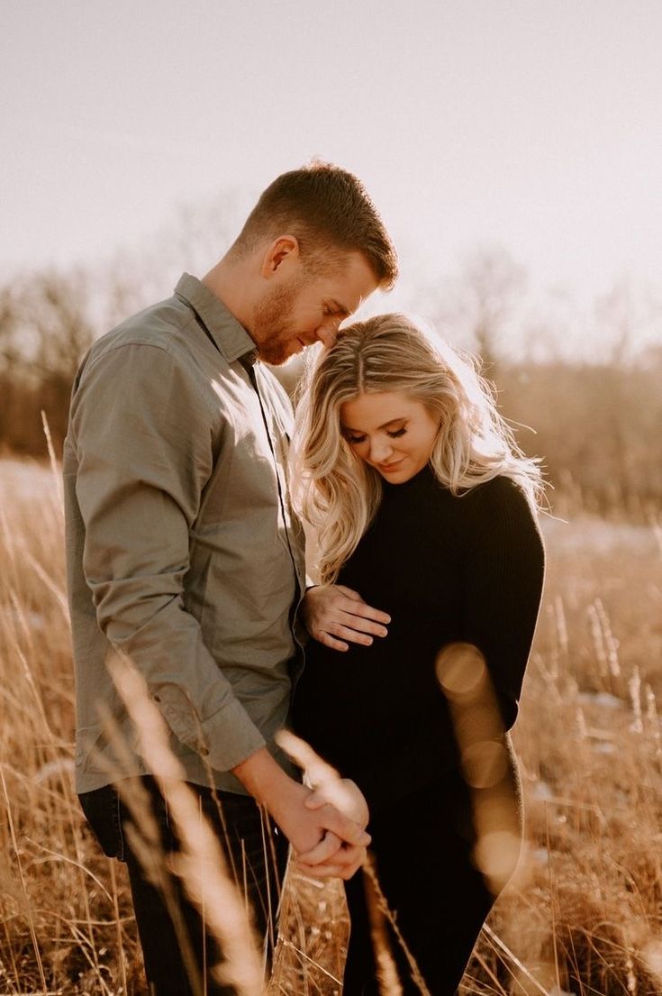 a man and woman standing in tall grass with their hands on each other's stomach