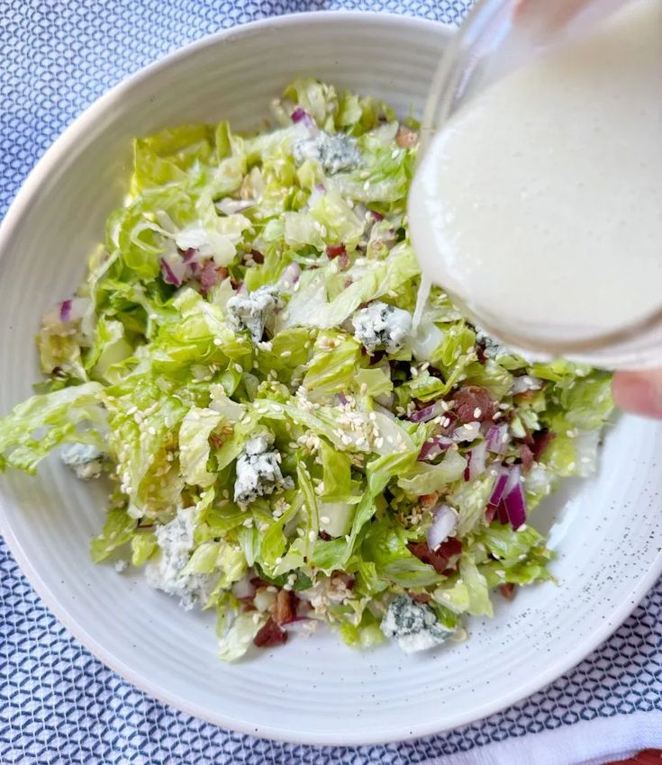 a person pouring dressing into a white bowl filled with lettuce and red onions