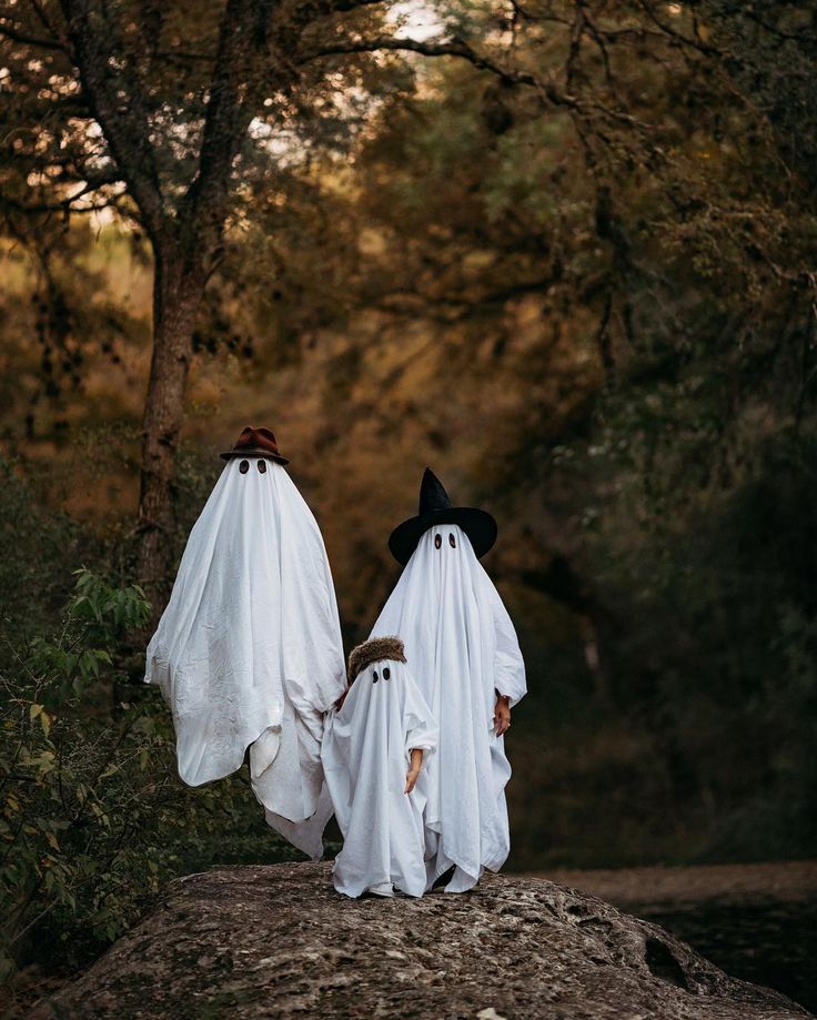 two people dressed in ghost costumes walking down a path through the woods with trees behind them