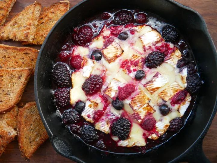 a skillet filled with fruit and cheese on top of a wooden table next to sliced bread