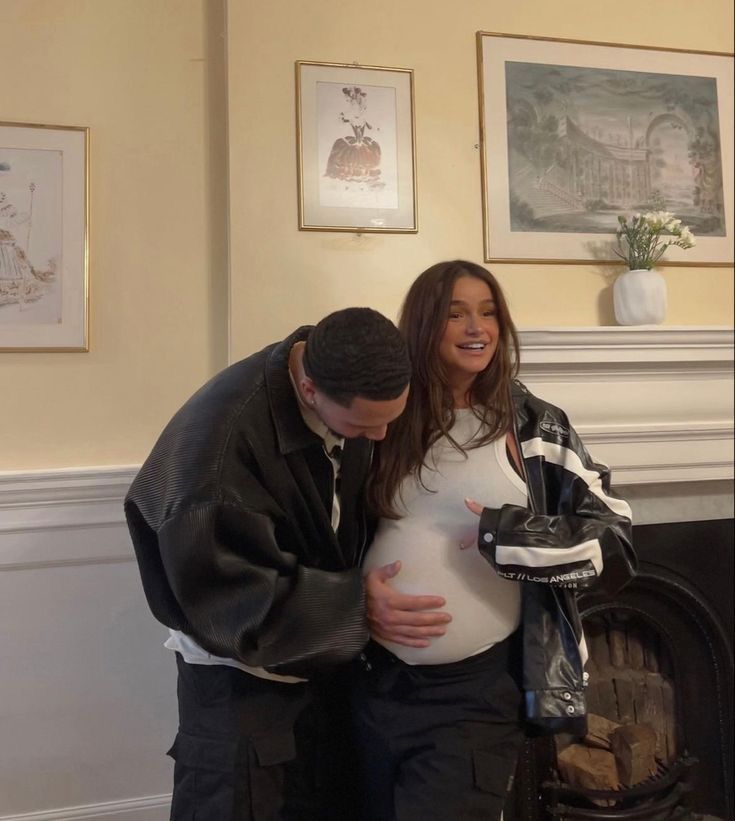 a man and woman standing next to each other in front of a fire place holding their pregnant belly