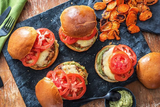 three burgers with tomatoes, lettuce and other toppings on a cutting board