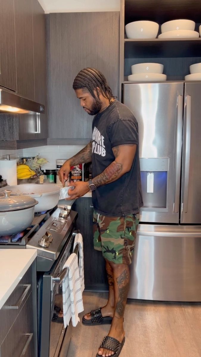 a man with dreadlocks standing in front of a stove preparing food on top of a pan