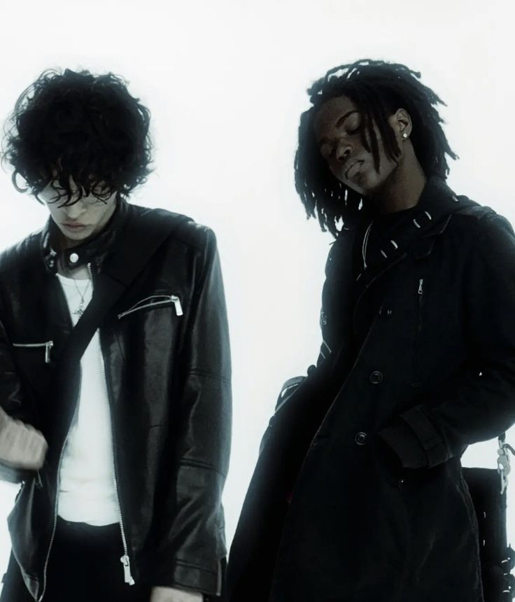 two young men standing next to each other in front of a white wall wearing black leather jackets